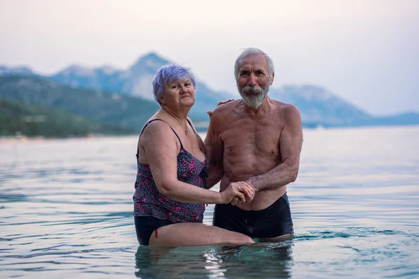 Elderly couple swims in the sea. Old bearded man and aged woman in swimsuits together stand in the tranqise water of Adriatic sea at sunset.