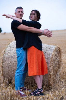 Genç bir çift, kuru saman balyalarının yakınındaki tarlada eğleniyor. Siyah tişörtlü ve kot pantolonlu bir adam siyah tişörtlü ve turuncu elbiseli bir kızla saman balyasının yanında dans ediyor..