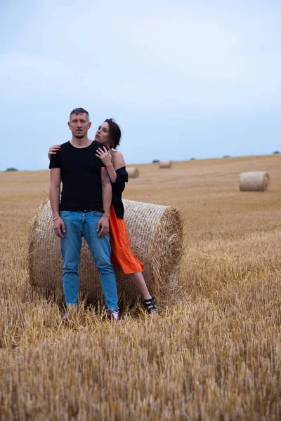 A young couple of beautiful people have fun in the field near round bales of dry hay. A man in a black T-shirt and jeans talks to a girl in a black shirt and an orange dress near a bale of straw.