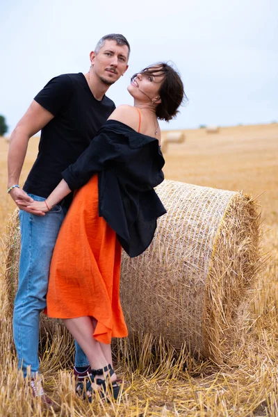A young couple of beautiful people have fun in the field near round bales of dry hay. A man in a black T-shirt and jeans dance with girl in a black shirt and an orange dress near a bale of straw.