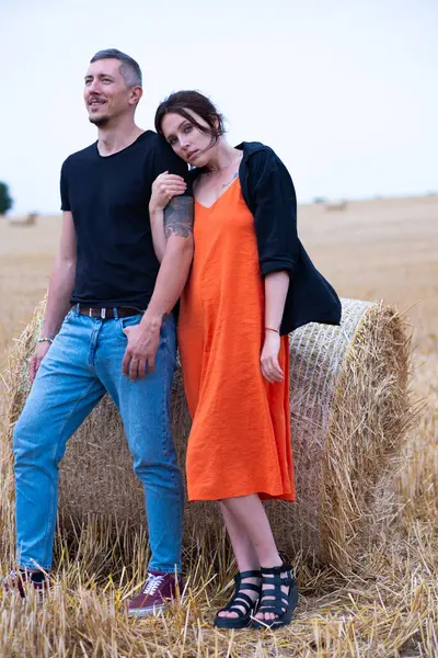 A young couple of beautiful people have fun in the field near round bales of dry hay. A man in a black T-shirt and jeans dance with girl in a black shirt and an orange dress near a bale of straw.
