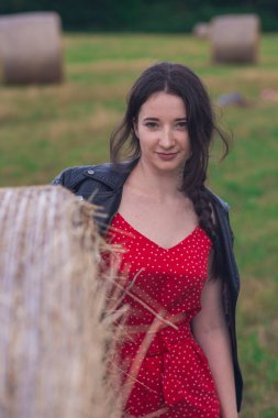 Avrupalı genç bir kız, sahada yuvarlak bir saman balyasının yanında güzel kırmızı bir elbise ve deri ceket giyiyor. Tarlanın arka planına, rüzgâr tarlalarına ve bulutlu gökyüzüne karşı..