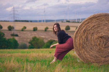 Sahadaki Bale of Straw 'da kırmızı pantolonlu ve siyah gömlekli genç Avrupalı Güzel Kız. Uzun saçlı genç kadın Bulutlu Gök ve Yel değirmenleri 'nin rüzgâr tarlasına karşı poz veriyor..