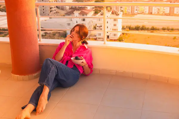 Beautiful girl on the balcony overlooking the sea. A girl in a red shirt and blue jeans rests on the balcony of her apartment on a high floor.