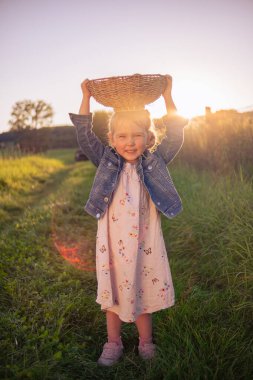 Üç yaşındaki güzel bir kız kafasında bir sepet elmayla poz veriyor. Güneşli bir çayırda kafasında elma sepeti olan küçük bir kız..