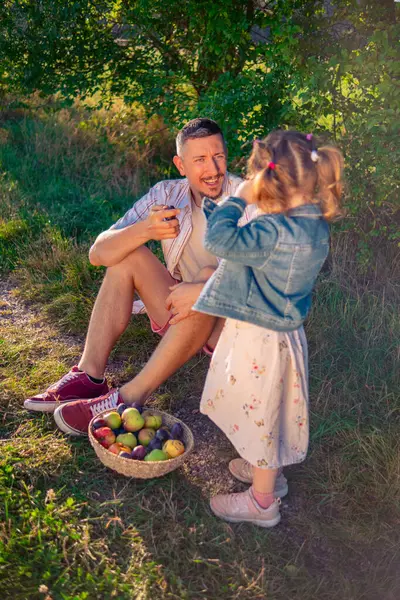 Küçük, güzel bir kız ve babası ağaçların gölgesinde otururken güneşte ıslanmış bir çayırda hasır sepetten elma yiyorlar..