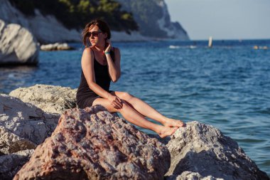 Kısa siyah bir elbise ve güneş gözlükleriyle denizin arka planında poz veren güzel bir kız. Deniz sahilinde zarif bir kadın..