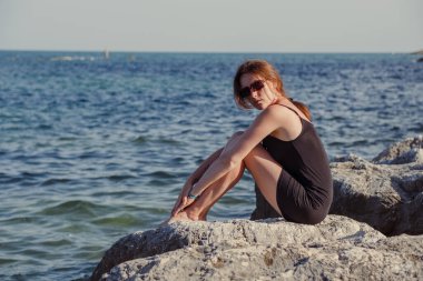 Kısa siyah bir elbise ve güneş gözlükleriyle denizin arka planında poz veren güzel bir kız. Deniz sahilinde zarif bir kadın..