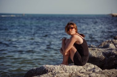 Kısa siyah bir elbise ve güneş gözlükleriyle denizin arka planında poz veren güzel bir kız. Deniz sahilinde zarif bir kadın..