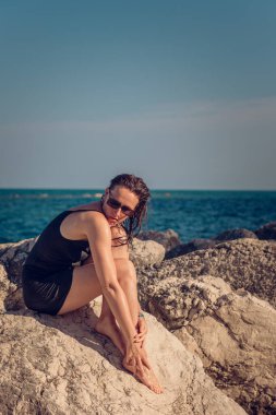 Kayalık bir sahilde kısa siyah bir elbise ve güneş gözlüğü takmış poz veren güzel bir kız. Zarif pozlar vermiş genç zarif bir kadın deniz kenarındaki büyük taşlarda duruyor..
