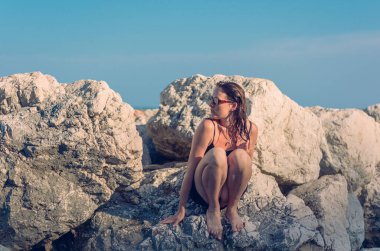 Kayalık bir sahilde kısa siyah bir elbise ve güneş gözlüğü takmış poz veren güzel bir kız. Zarif pozlar vermiş genç zarif bir kadın deniz kenarındaki büyük taşlarda duruyor..