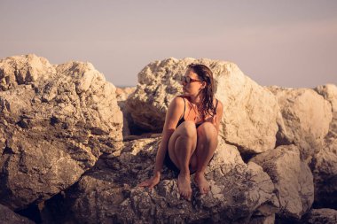 Kayalık bir sahilde kısa siyah bir elbise ve güneş gözlüğü takmış poz veren güzel bir kız. Zarif pozlar vermiş genç zarif bir kadın deniz kenarındaki büyük taşlarda duruyor..