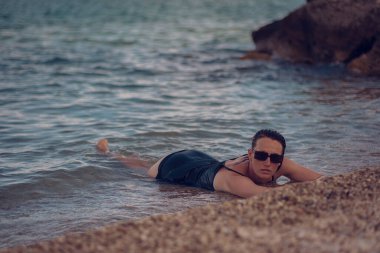 Deniz sahilinde kısa siyah bir elbise ve güneş gözlükleriyle poz veren güzel bir kız. Denizde poz veren zarif genç bir kadın..