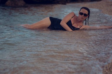 Deniz sahilinde kısa siyah bir elbise ve güneş gözlükleriyle poz veren güzel bir kız. Denizde poz veren zarif genç bir kadın..