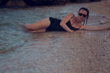 Deniz sahilinde kısa siyah bir elbise ve güneş gözlükleriyle poz veren güzel bir kız. Denizde poz veren zarif genç bir kadın..