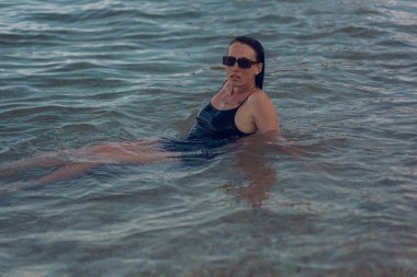 Deniz sahilinde kısa siyah bir elbise ve güneş gözlükleriyle poz veren güzel bir kız. Denizde poz veren zarif genç bir kadın..