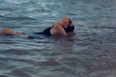 Deniz sahilinde kısa siyah bir elbise ve güneş gözlükleriyle poz veren güzel bir kız. Denizde poz veren zarif genç bir kadın..