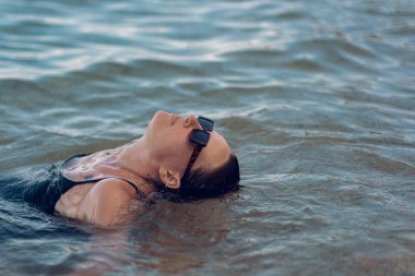 Deniz sahilinde kısa siyah bir elbise ve güneş gözlükleriyle poz veren güzel bir kız. Denizde poz veren zarif genç bir kadın..
