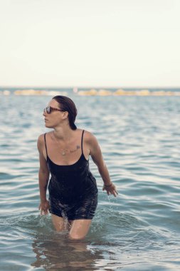 Deniz sahilinde kısa siyah bir elbise ve güneş gözlükleriyle poz veren güzel bir kız. Denizde poz veren zarif genç bir kadın..