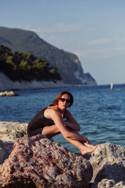 Kısa siyah bir elbise ve güneş gözlükleriyle denizin arka planında poz veren güzel bir kız. Deniz sahilinde zarif bir kadın..