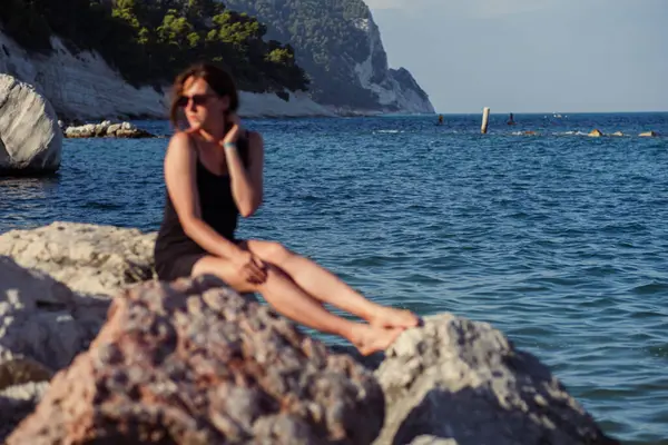 Kısa siyah bir elbise ve güneş gözlükleriyle denizin arka planında poz veren güzel bir kız. Deniz sahilinde zarif bir kadın..