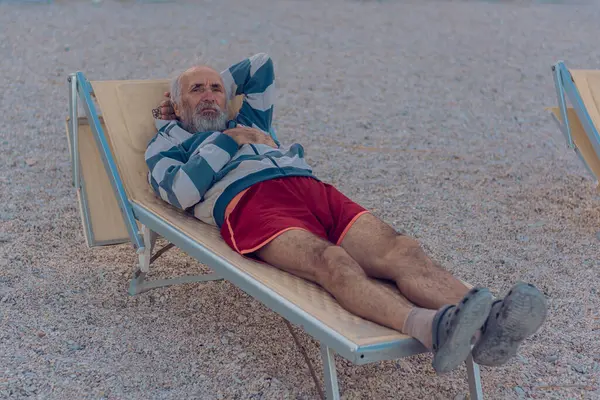 An old man is lying on a sun lounger on the beach. An elderly gray-haired tourist with a beard in red shorts is enjoying himself on the beach by the sea. Grandfather alone on the sea beach