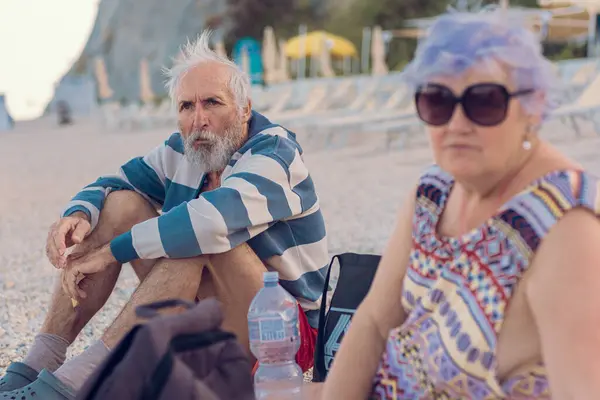 Grandfather and grandmother are sitting by the sea. A colorful couple of old people are relaxing on the beach. Old tourists enjoying life