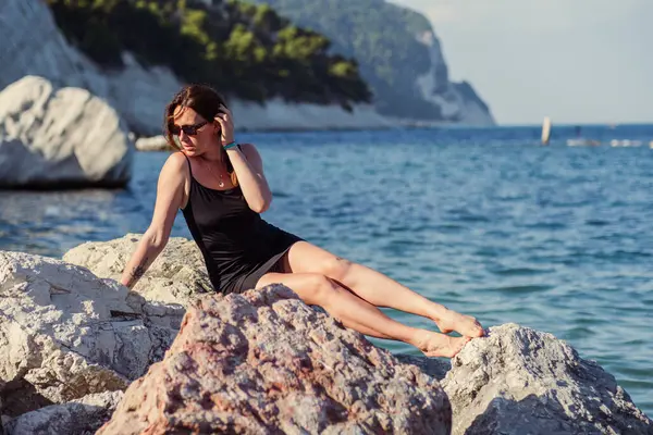 Kısa siyah bir elbise ve güneş gözlükleriyle denizin arka planında poz veren güzel bir kız. Deniz sahilinde zarif bir kadın..