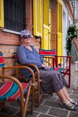 Yaşlı bir kadın sarı panjurlu tuğla bir evin duvarının yanında renkli bir sandalyede oturuyor. Güneş gözlüklü bir kadın, güney şehrinde bir sandalyede otururken dinlenmekten zevk alır..