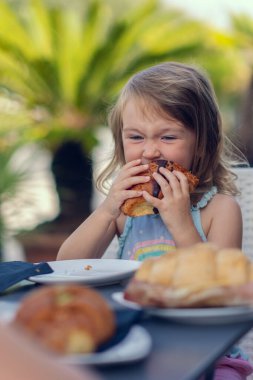 A beautiful little girl eats a croissant with chocolate with appetite. The girl has breakfast with a croissant. Caucasian girl eats pastries. Close-up photo.