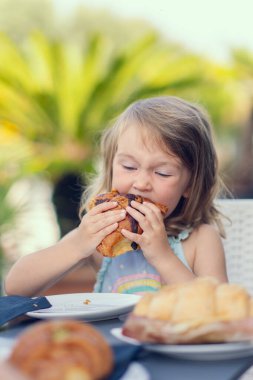 A beautiful little girl eats a croissant with chocolate with appetite. The girl has breakfast with a croissant. Caucasian girl eats pastries. Close-up photo.