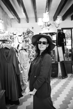 Photography in vintage style in black and white. A young girl tries on a hat in a store. Inside the boutique there is a characteristic atmosphere - a lot of clothes and accessories.