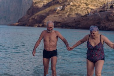 Old people swim in the sea. An elderly couple enjoys life by spending time at sea. An old grandfather and grandmother in a swimsuit are walking along the sea.