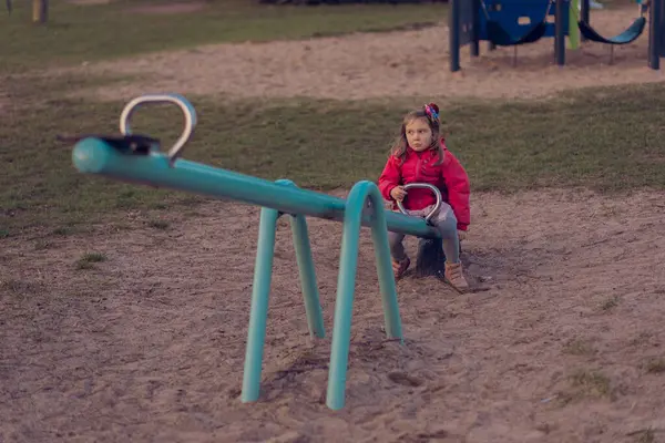 stock image A beautiful little girl plays alone on the playground. The girl is bored alone and has no one to ride with on the teeter-totter or seesaw and she sits on one side of the swing.