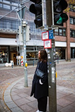 Bir Alman şehrinin sonbahar günü kavşağında elinde çantasıyla trafik ışığında duran siyah paltolu bir kız. Genç bir kadın yeşil ışık için bekliyor. Şehir hayatının doğal gerçekçi fotoğrafı..