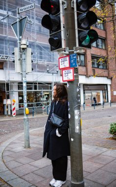 Bir Alman şehrinin sonbahar günü kavşağında elinde çantasıyla trafik ışığında duran siyah paltolu bir kız. Genç bir kadın yeşil ışık için bekliyor. Şehir hayatının doğal gerçekçi fotoğrafı..