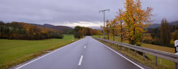 stock image Wide autumn panorama of asphalt road and trees on sides. Yellowed trees and still green grass, a smooth asphalt road and bumper on the side.