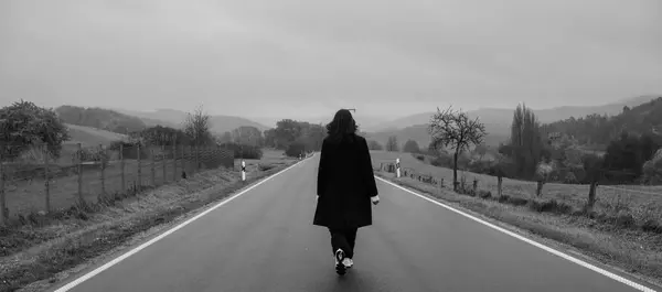 Siyah ceketli genç bir kadın, sarı ağaçlar ve çimlerle çevrili asfalt bir yolda tek başına yürüyor. Dramatik gökyüzü ve arka planda dağlar. Siyah ve beyaz resim.