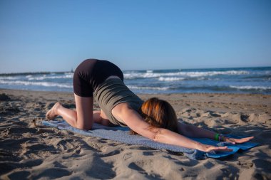 Spor kıyafetli kız deniz kumsalında yoga yapıyor. Açık mavi gökyüzünün altında deniz kıyısında sabah yogası. Sportif genç kadın sağlıklı yaşam tarzını sürdürüyor.