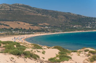 Bir dağın altında uzanan vahşi bir plajın güzel manzarası. Bir sürü tatilciyle, sörfçüyle, rüzgar sörfçüsüyle. İspanya 'da deniz kıyısında büyük bir kumsal manzarası.