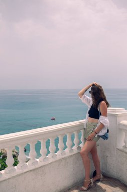 Denize bakan bir balkonda poz veren güzel bir kız. Güneş gözlüklü ve beyaz bluzlu bir kız İspanya 'da deniz kenarında tatilin tadını çıkarıyor. Kadife mevsiminde denizde tatil.