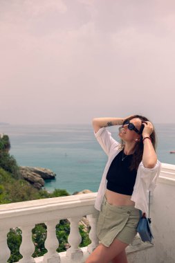 Denize bakan bir balkonda poz veren güzel bir kız. Güneş gözlüklü ve beyaz bluzlu bir kız İspanya 'da deniz kenarında tatilin tadını çıkarıyor. Kadife mevsiminde denizde tatil.