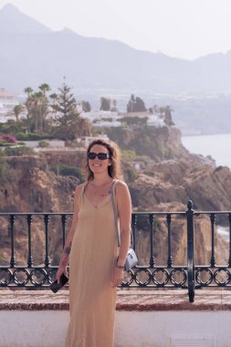 Beautiful girl posing on a balcony overlooking the sea. Girl in sunglasses and a long dress enjoying a seaside vacation in Spain. Vacation at sea in the velvet season.