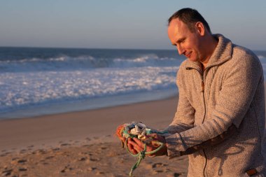 Bir adam plajda toplanan büyük bir plastik atık yığınını tutuyor. Genç bir kadın, okyanus tarafından sahile atılan çeşitli plastik atıkları tutuyor..