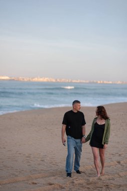 Okyanus sahilinde poz veren genç bir çift. Dalgalar ve ufukta bir şehir. Plajda yürüyen sağlıklı ve güzel Avrupalı erkek ve kadın. Arka planda okyanus olan genç bir çift..
