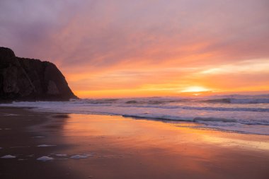 Okyanus dalgaları büyüleyici bir günbatımının altında karanlık kumlu bir plaja yavaşça yuvarlanıyor. Gökyüzü, ıslak kuma yansıyan turuncu, pembe ve mor tonlarda boyanmıştır. Huzurlu ve rahatlatıcı bir atmosfer, seyahat, doğa ve sakinleştirici kavramları için mükemmel.