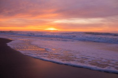 Okyanus dalgaları büyüleyici bir günbatımının altında karanlık kumlu bir plaja yavaşça yuvarlanıyor. Gökyüzü, ıslak kuma yansıyan turuncu, pembe ve mor tonlarda boyanmıştır. Huzurlu ve rahatlatıcı bir atmosfer, seyahat, doğa ve sakinleştirici kavramları için mükemmel.