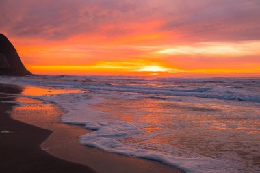Okyanus dalgaları büyüleyici bir günbatımının altında karanlık kumlu bir plaja yavaşça yuvarlanıyor. Gökyüzü, ıslak kuma yansıyan turuncu, pembe ve mor tonlarda boyanmıştır. Huzurlu ve rahatlatıcı bir atmosfer, seyahat, doğa ve sakinleştirici kavramları için mükemmel.
