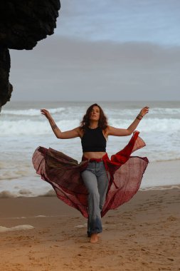 Hint tarzı kırmızı etekli güzel bir kadın okyanus kıyısında kayaların yanında poz veriyor. Sahilde pareo dansı yapan şık bir kız, şık ve şık bir sanat fotoğrafçısı..