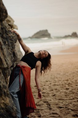 Hint tarzı kırmızı etekli güzel bir kadın okyanus kıyısında kayaların yanında poz veriyor. Sahilde pareo giymiş şık bir kız, şık ve şık bir sanat fotoğrafçısı..